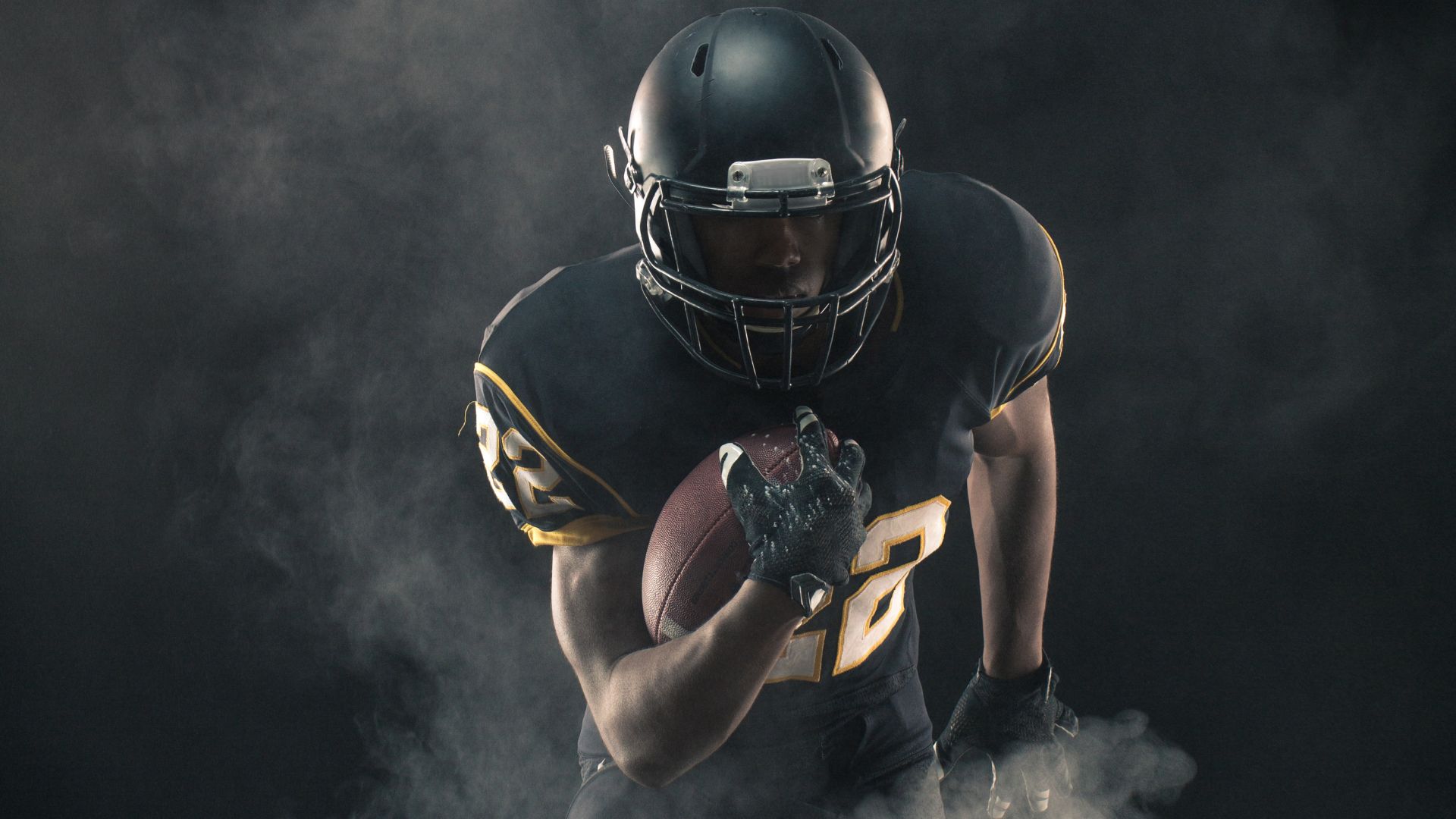 African American football player dressed in full uniform holding football in front of a black backdrop with smoke rising from the ground.