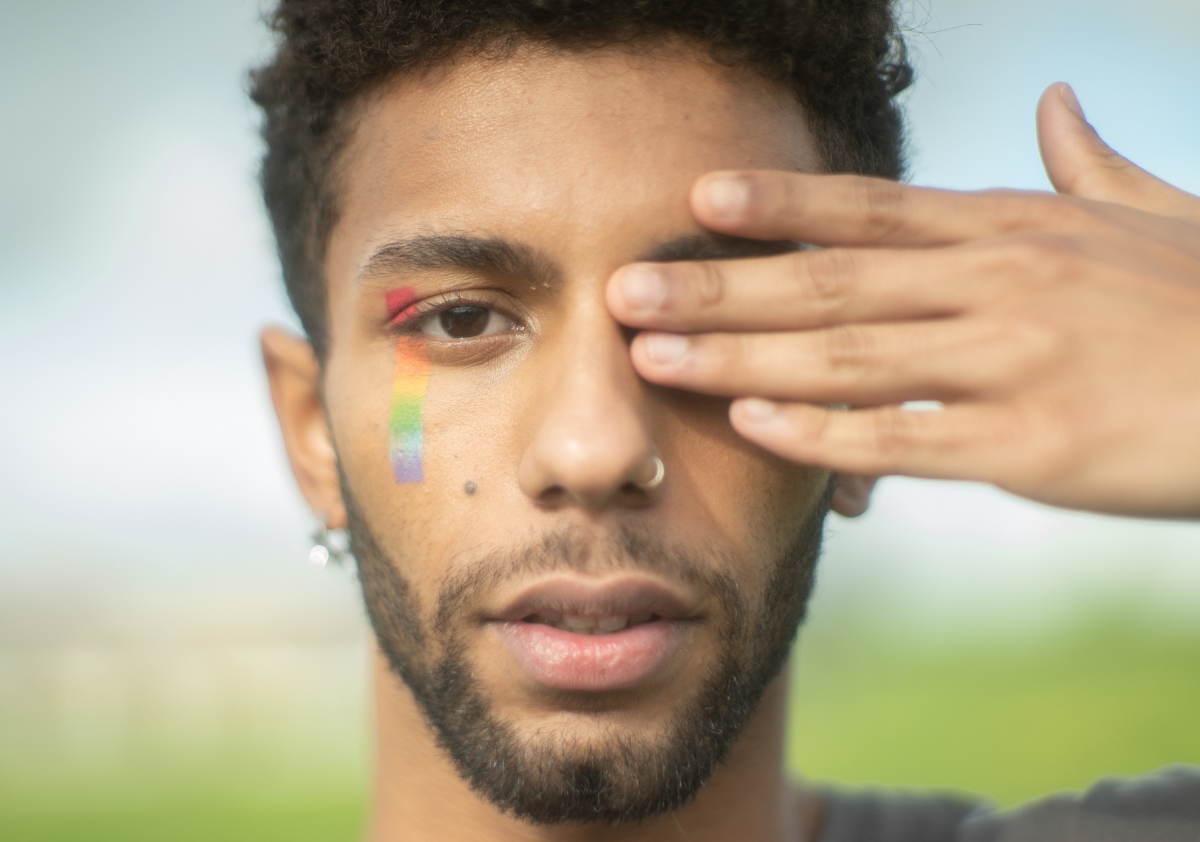 Young man of color with hand covering his eye and rainbow makeup on his face in alignment with community building during pride month