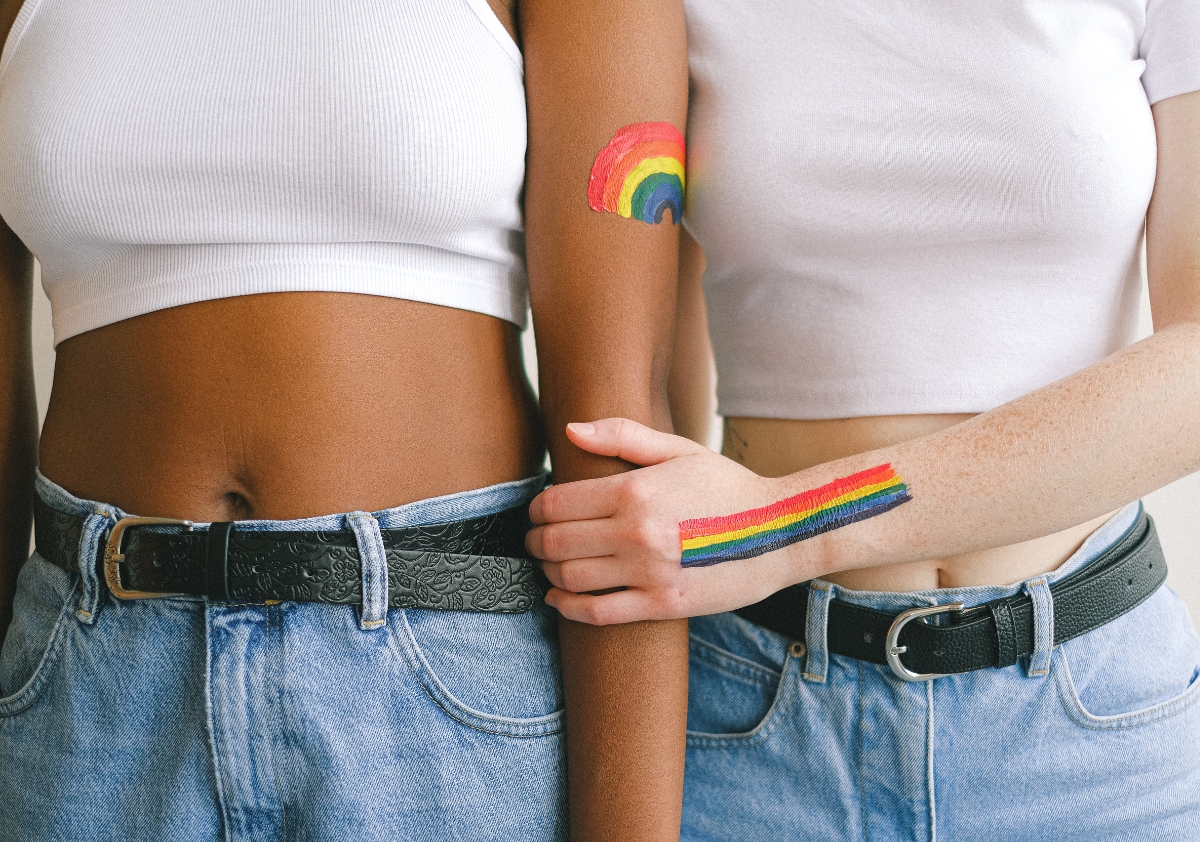 Two women from the chest down wearing white tank tops and denim jeans adorned with rainbow flags painted on their arms in alignment with building community during Pride.