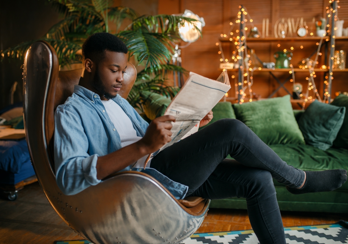 A young Black male is sitting down in a swanky coffee shop reading a newspaper that prioritizes diversity in media