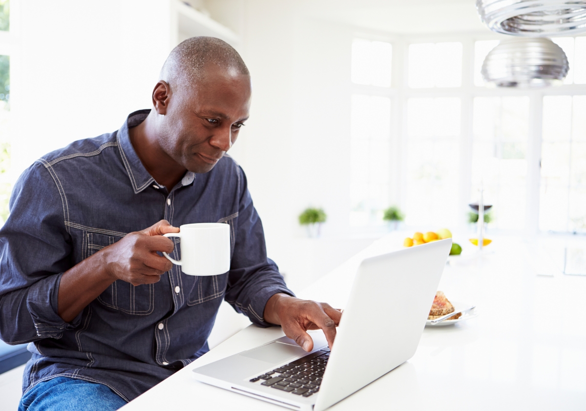 A black man sits at his dining room table sipping coffee while enjoying remote work and great company culture.