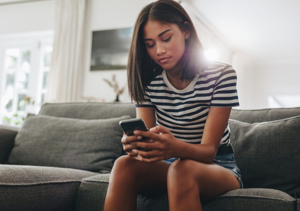 Young woman of color sits in her living room scrolling through her phone browsing innovative marketing ideas from brand