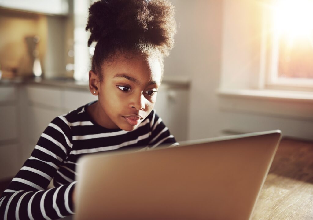 Young Black girl with beautiful afro pulled into a high puff sits in her kitchen browsing innovative marketing ideas from a  brand on her computer
