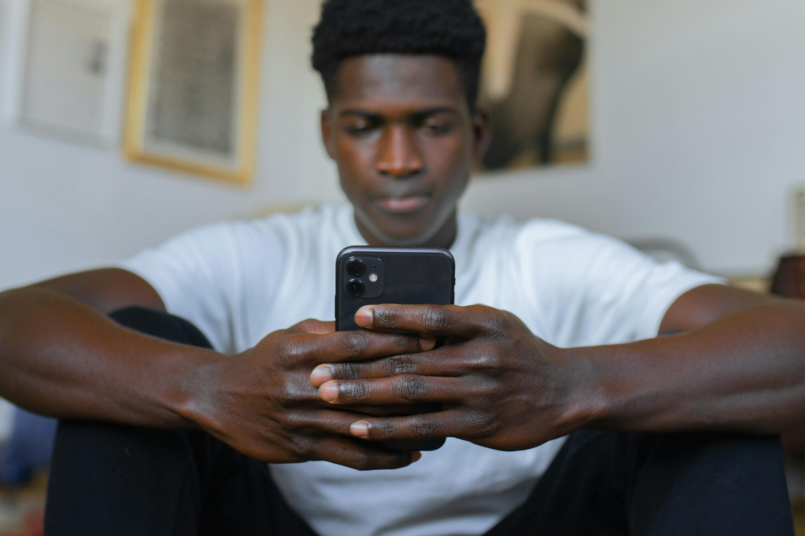 A gen z young Black man with a focused expression is seated indoors, wearing a white shirt and holding a smartphone in both hands. The background is softly blurred, featuring framed artwork on the wall, emphasizing his engagement with the phone.